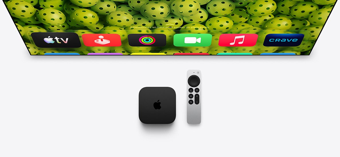 Une Apple TV 4K et une télécommande Siri Remote placées sous un écran de télévision. L’écran d’accueil affiche la rangée supérieure des icônes d’apps, avec des balles de pickleball vertes en arrière-plan