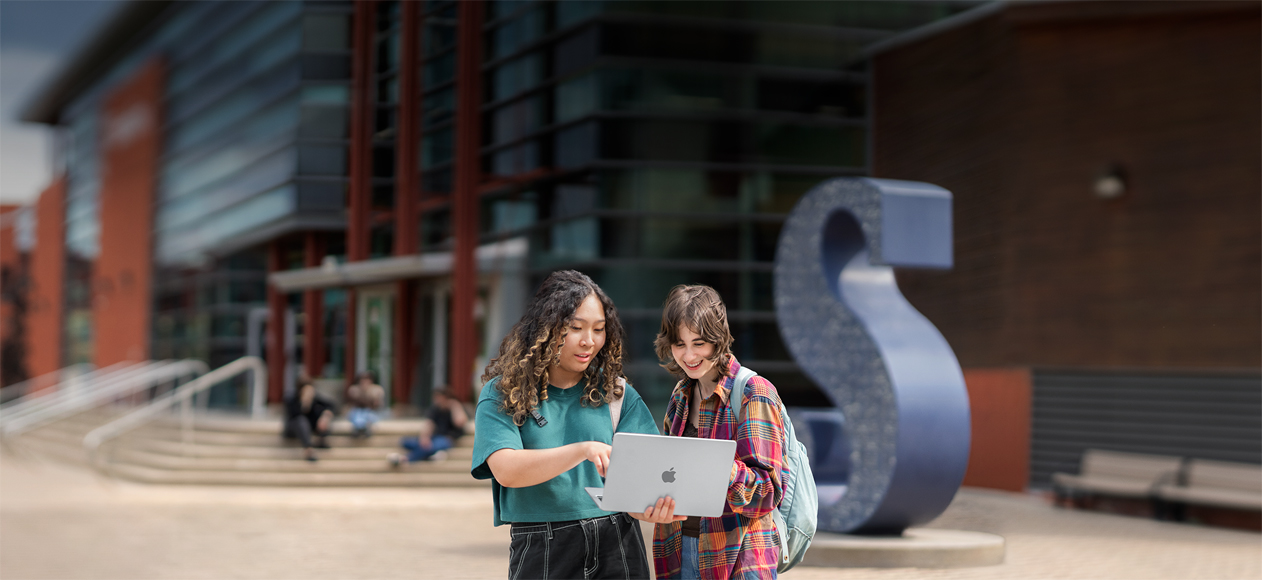Deux personnes sont debout dehors sur le campus de leur établissement scolaire, l’une d’entre elles tient un MacBook avec lequel elles interagissent toutes les deux en souriant