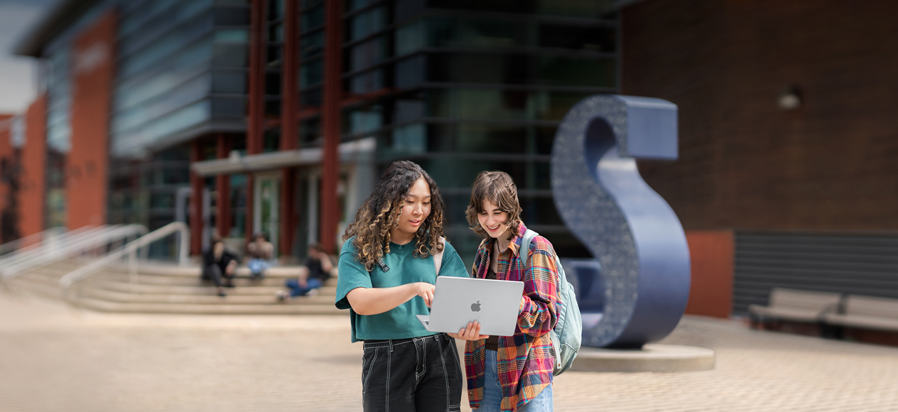 Two students stand outside on campus, one of them holding a MacBook that they're both engaged with, smiling