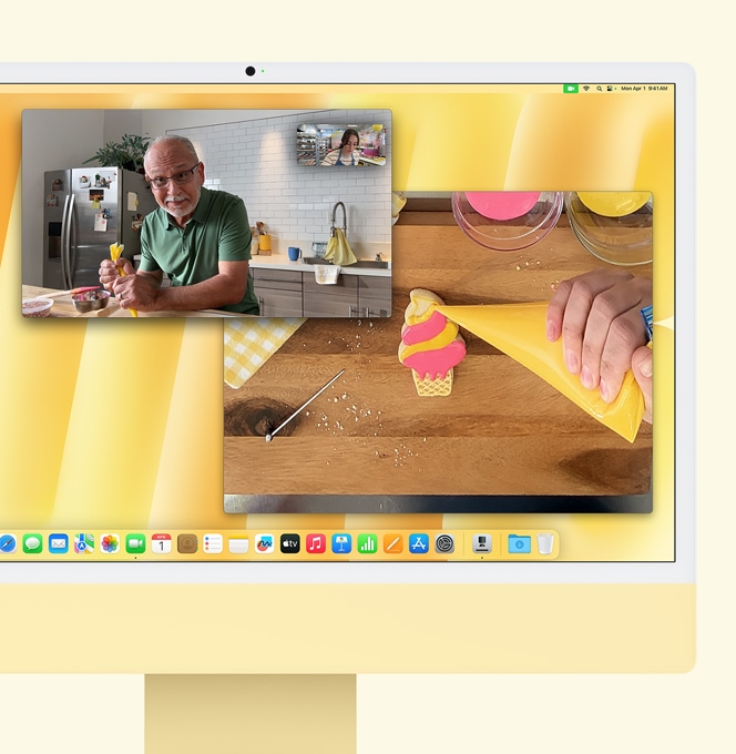 Žuti iMac. Demonstracija prikaza radnog stola. Na zaslonu se prikazuje videopoziv između slastičarke i još jedne osobe. Na jednom se zaslonu prikazuje slastičarka i lice muškarca. Na drugom se zaslonu prikazuje kako slastičarka nanosi glazuru na kolačić