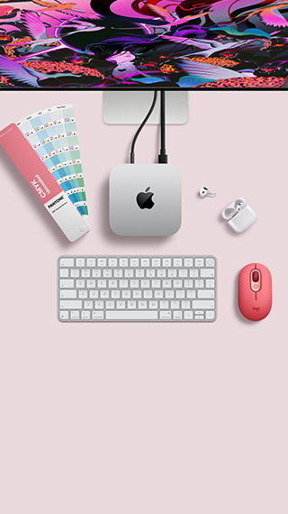Skats no augšas uz Mac mini sudraba krāsas alumīnija korpusā ar noapaļotiem stūriem un spīdīgu melnu Apple logotipu centrā, savienots ar vadu. Augšpusē daļēji redzams Studio Display, atvērti AirPod 4, Pantone krāsu paletes, cipartastatūra un pele