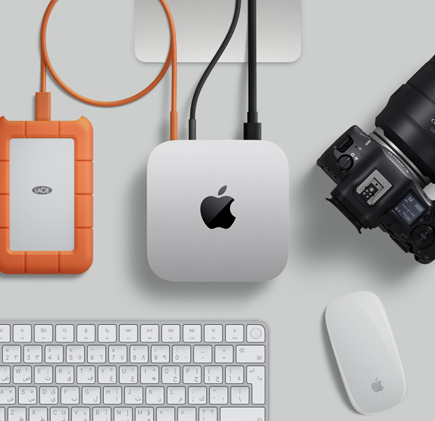 لقطة علوية لجهاز Mac&nbsp;mini فضي اللون، مع سلكين متصلين من المنافذ الخلفية بشاشة، وبجواره ماجيك ماوس أسود ولوحة مفاتيح ماجيك سوداء