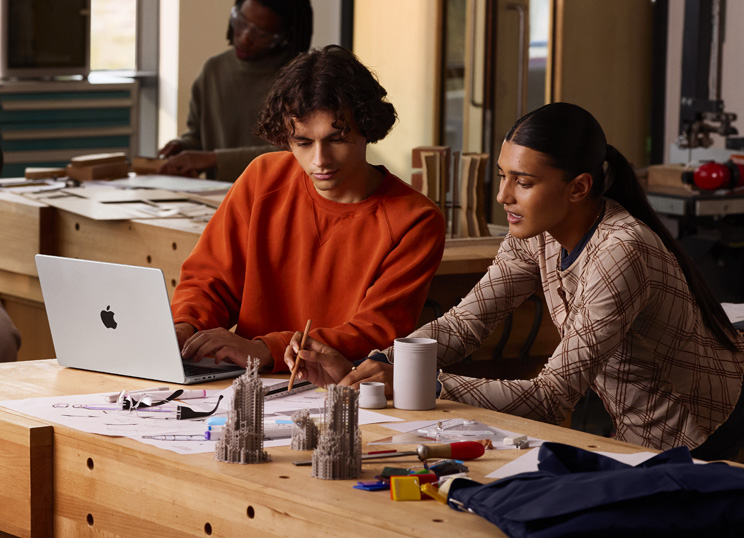 Dos personas colaboran en un proyecto en una mesa, una escribe en una MacBook, la otra usa un lápiz y una regla sobre un papel, hay materiales de arte alrededor.