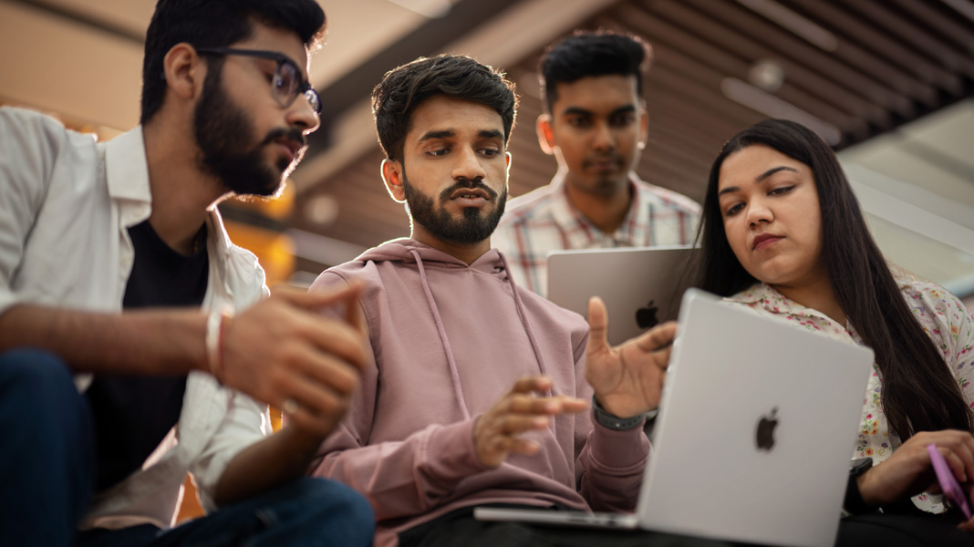 A group of four students gathered around a MacBook engaged in collaborative discussion.