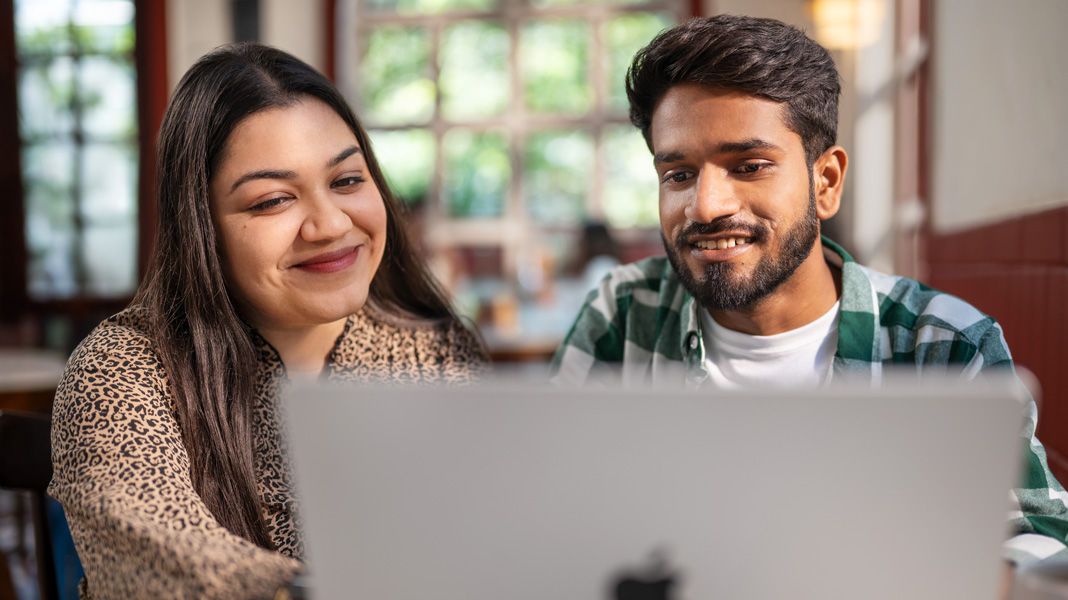 Two students smile warmly while looking at an open MacBook in a bright, airy indoor setting.