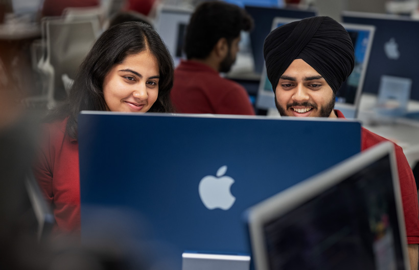 Two smiling students in a classroom look at their shared iMac screen.