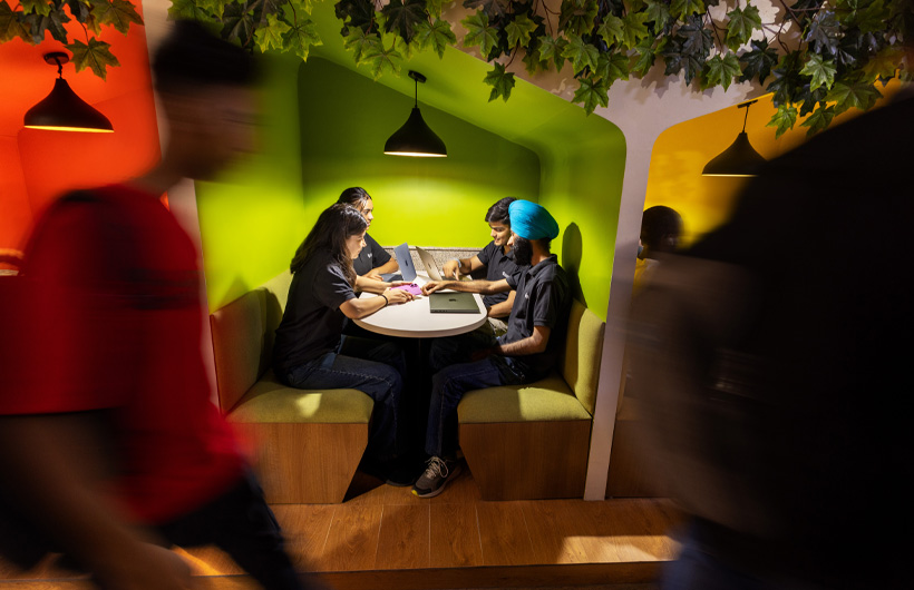 In a modern space with colorful lighting, four students at a booth work in pairs on their shared MacBook.
