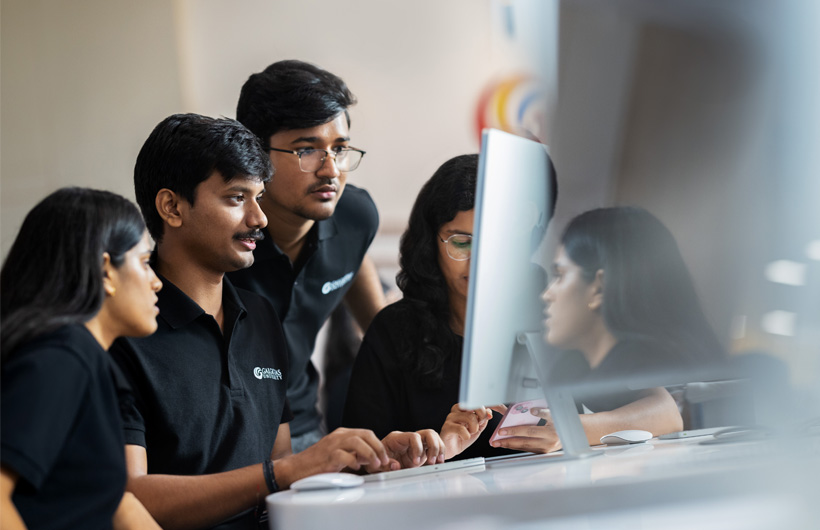 An instructor sits at an iMac while his students huddle around, watching him work.