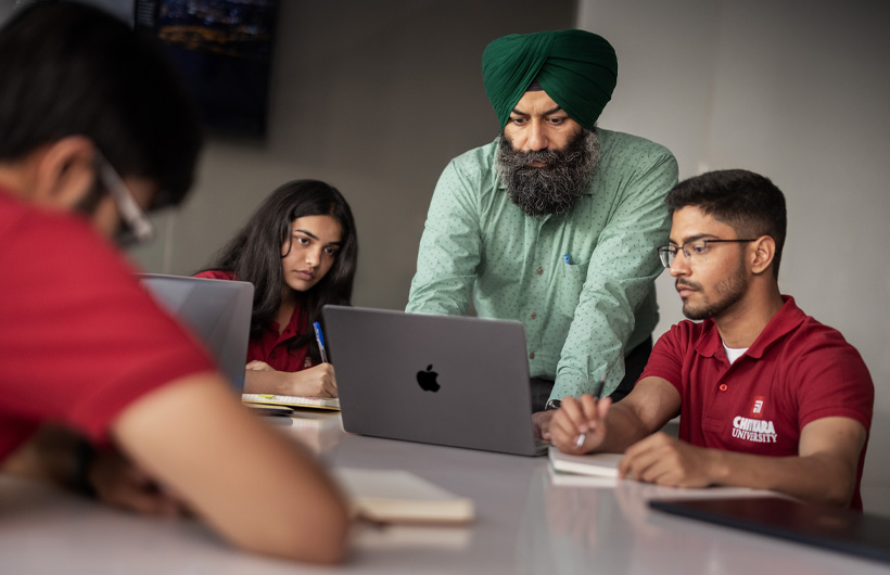 A professor at a classroom table leans over to look at his students’ MacBook screen.