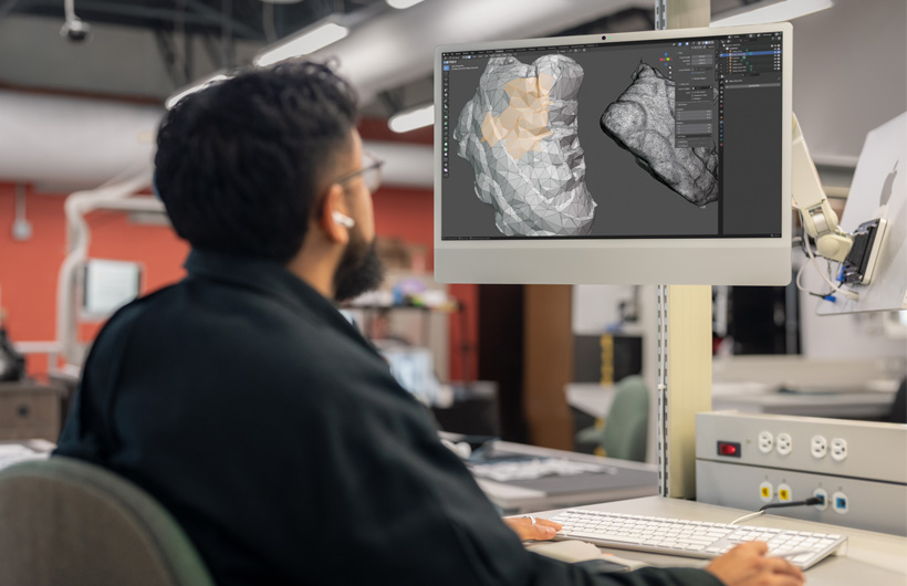 A student sits at a desk reviewing two 3D-scanned head models displayed side by side on iMac monitors.