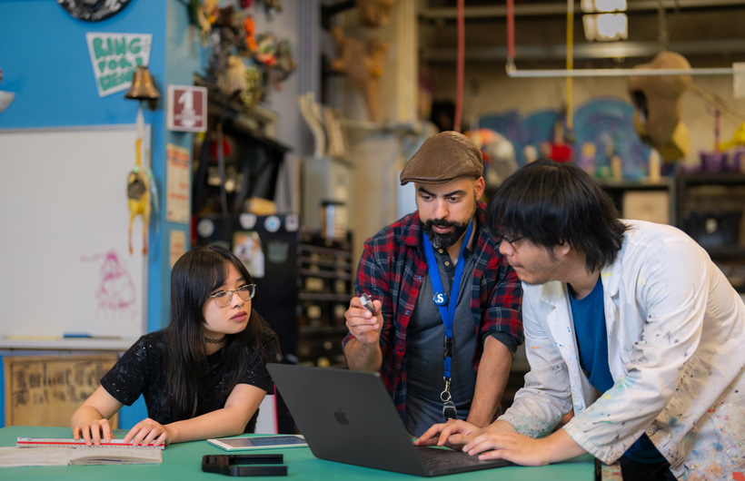 Three students huddle around a MacBook sitting on a table in a cluttered, colorful workshop filled with tools and artwork.